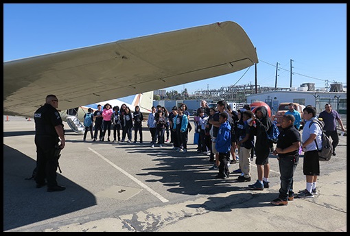 Middle school students standing near a small plane as they are being letured by a K-9 team (officer and dog).