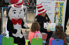 A police officer in a 'cat in the hat' hat while reading to children, the officer is also sitting next to a cat in the hat character.